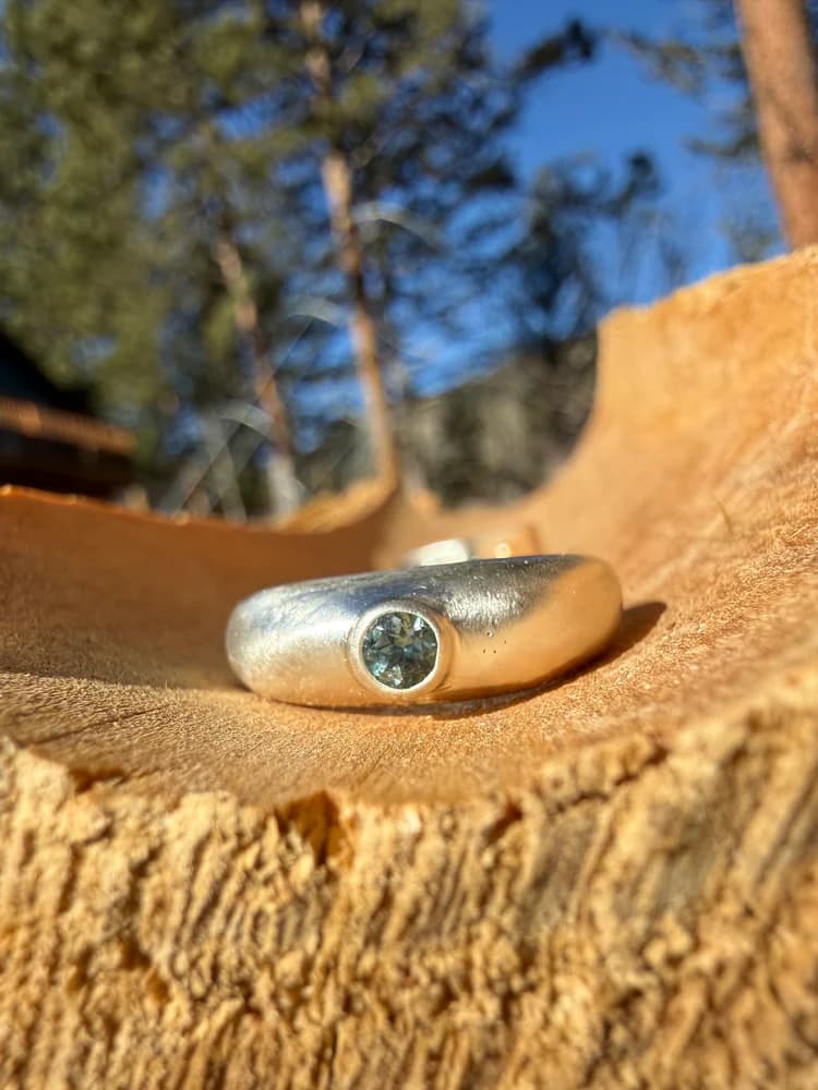 Handmade silver ring with a round teal stone, resting in weathered wood with trees and sky behind