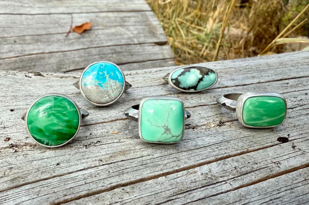 Five statement silver rings with green and blue stones on a weathered wooden log