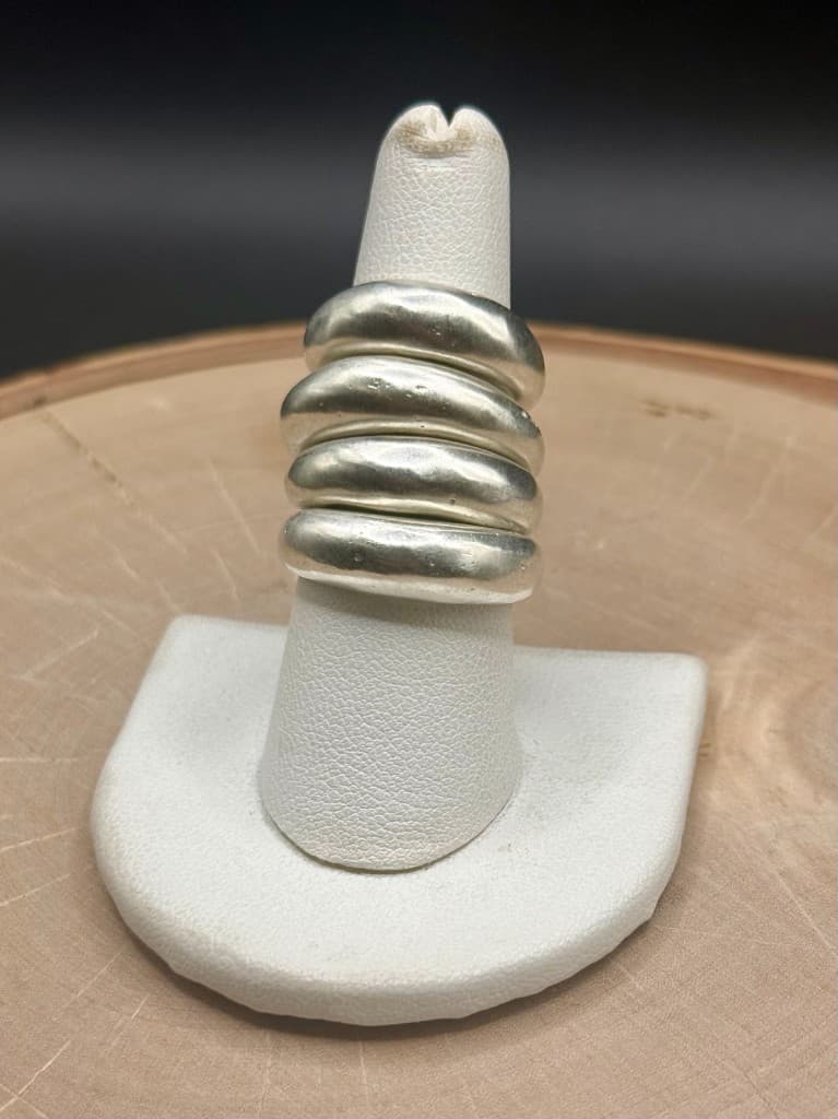 Four chunky brushed silver rings stacked on a white ring mandrel on a wood slice against a dark background