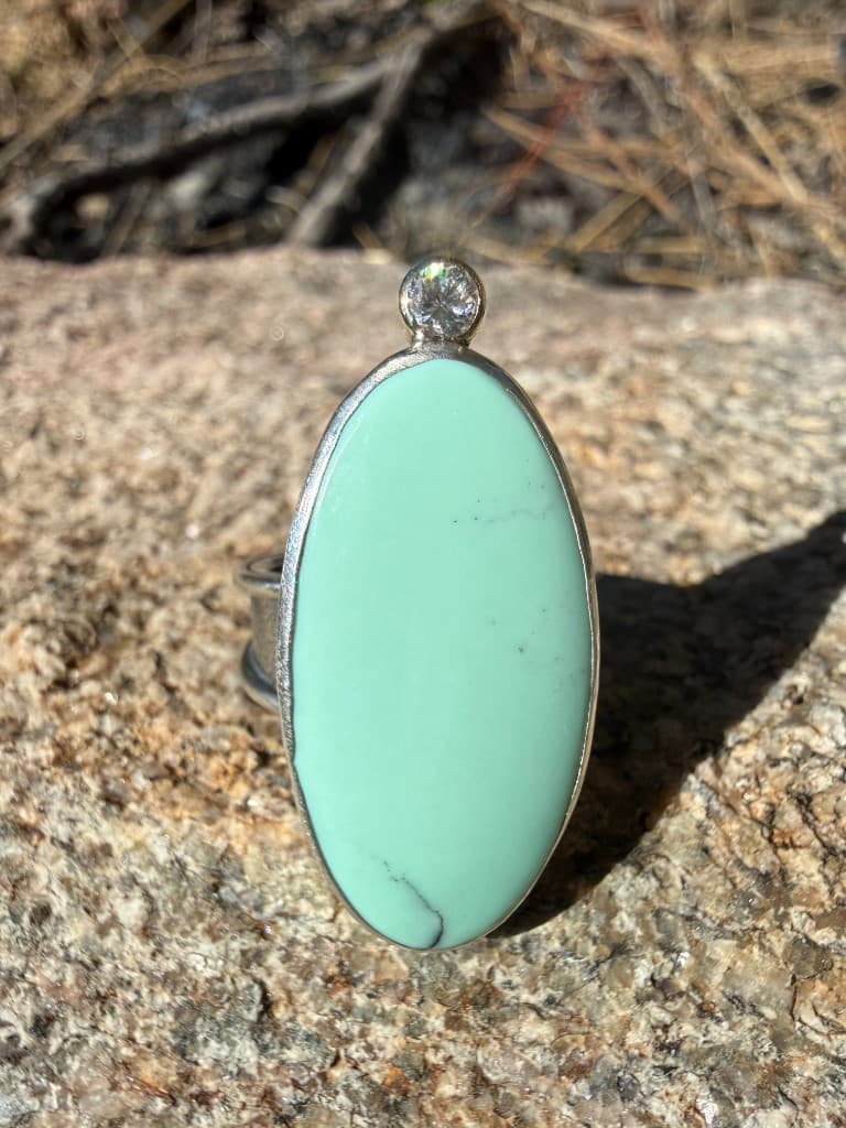 Handmade silver ring with a large oval mint-green stone and a small faceted accent, resting on textured rock in sunlight