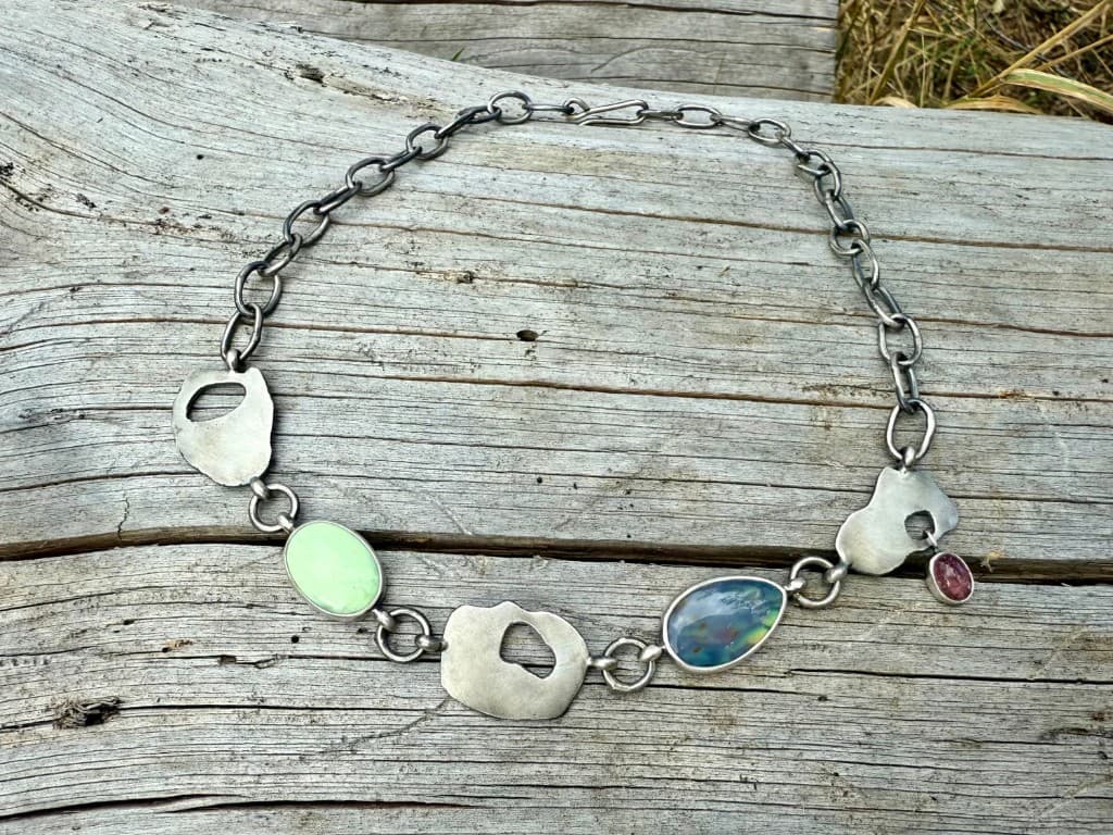 Sterling silver necklace with seafoam, teal, and rose-toned stones in bezels, arranged on weathered wood outdoors