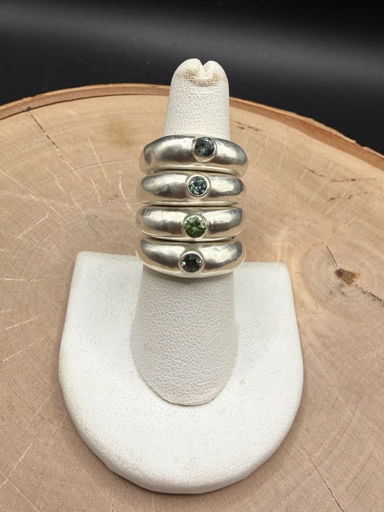 Four chunky brushed silver rings with teal and green gemstones on a white display stand