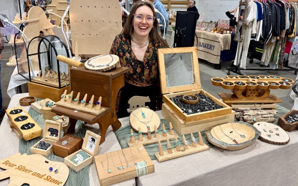 Bear Silver & Stone booth at a craft market — jewelry on wooden displays with a carved bear sign, Claire behind the table